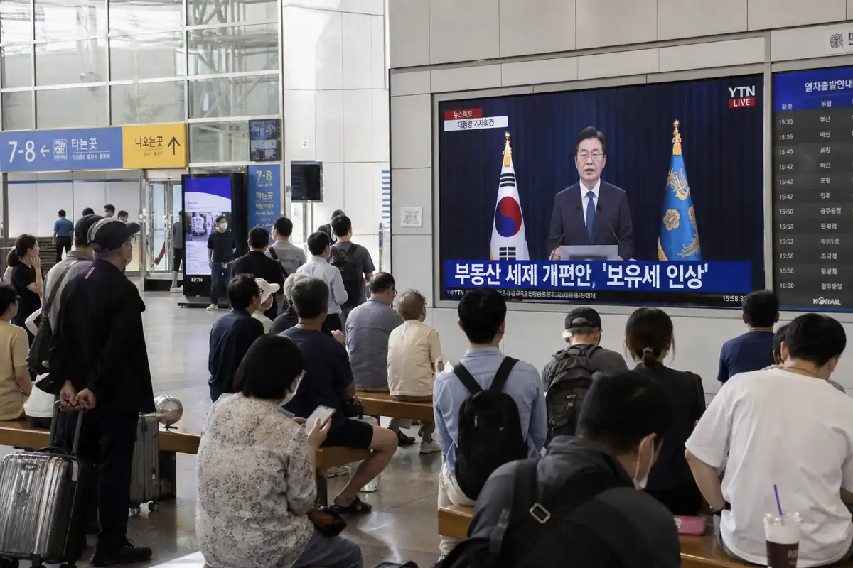 2026년 대한민국 서울역 대합실 TV 화면에 부동산 세제 개편안 보유세 인상을 발표하는 대통령의 취임 1주년 기자회견 생중계가 방송되고 있는 실제 현장 사진